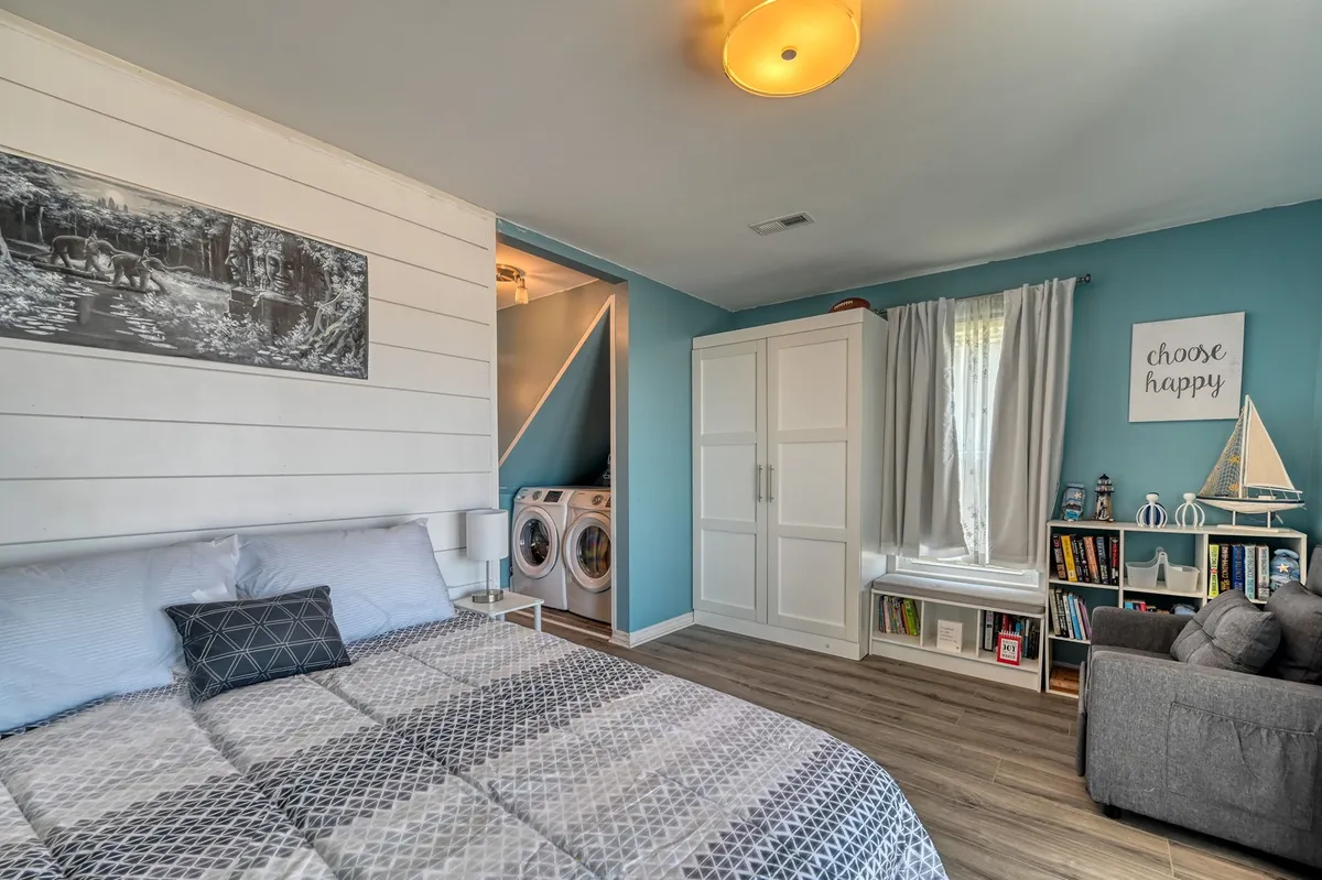 This image showcases a bright, cheerful bedroom with patterned bedding, a nearby laundry area, decorative wall art, and a cozy reading nook tucked in the corner. The room feels welcoming and playful, suitable for kids to recharge or enjoy quiet reading time.