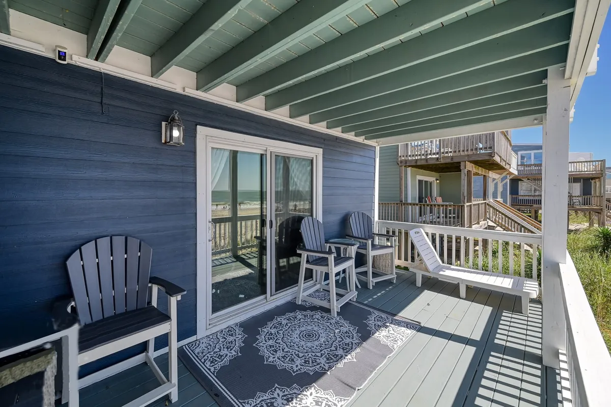 A screened porch with patio chairs, a decorative rug, a sliding glass door, and a tranquil view of the beach. The space is shaded and breezy, providing a comfortable spot for parents to relax, kids to snack, and pets to nap after beach outings.