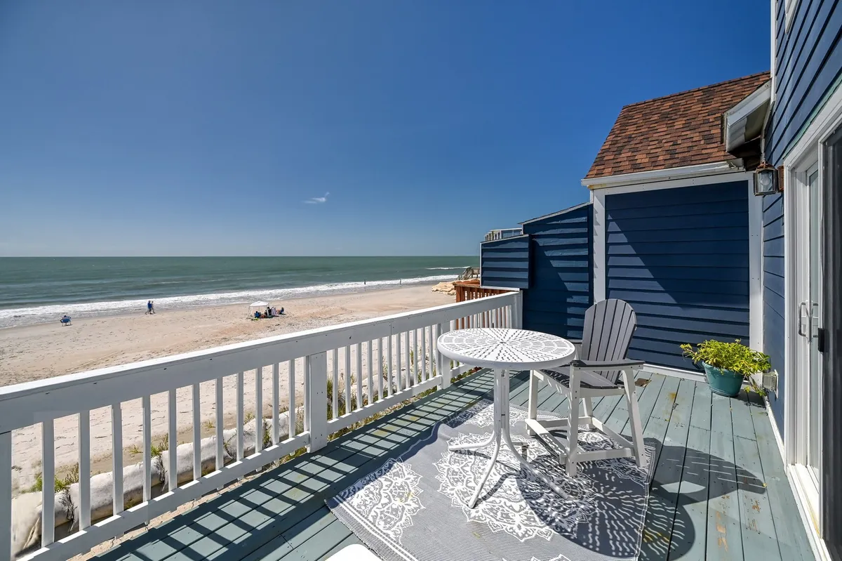 An inviting outdoor table and chairs set on a deck facing the beach and sea, with a decorative rug and potted plant adding extra charm. The view stretches out to the water, creating an ideal spot for reading, relaxing, or sharing a meal while taking in the ocean air.