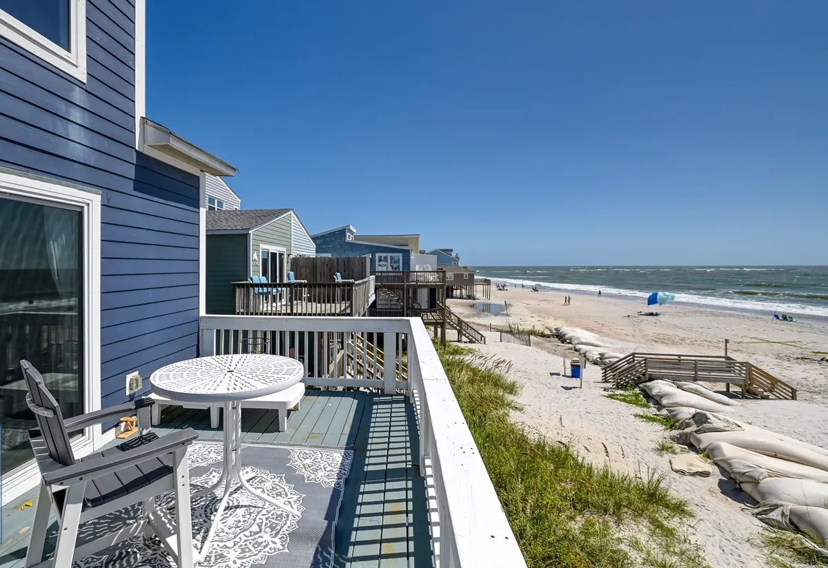 A serene deck overlooking the beach, complete with a table and chairs ideal for outdoor meals or relaxing as waves roll in. The view showcases soft sand, gentle waves, and neighboring coastal homes, inviting gatherings and quiet moments alike.