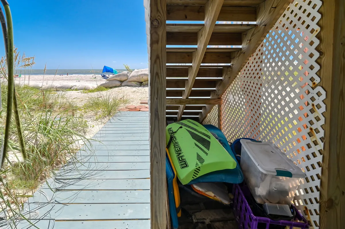 A wooden boardwalk stretches from the property directly onto the sandy beach, flanked by colorful beach gear and framed by the dunes. Wooden steps lead down to the shore, and the scene is bathed in inviting sunlight, evoking a laid-back, family-friendly vibe.