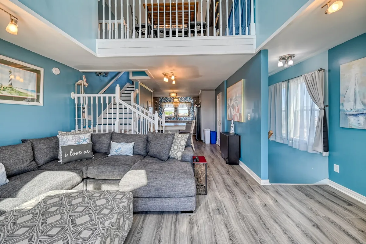 A spacious sectional sofa faces a light-filled dining area, accented by a staircase leading to the upper level and decorative wall art. The setting is casual but comfortable, perfect for group relaxation, shared meals, or evening wind-downs after a day outside.
