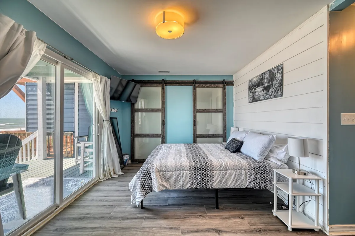 A tranquil bedroom with a large bed, sunlight streaming in through expansive windows, decorative wall art, and sliding barn doors. The room has a peaceful, inviting mood and looks as though it offers private access to a deck or an impressive outdoor view.