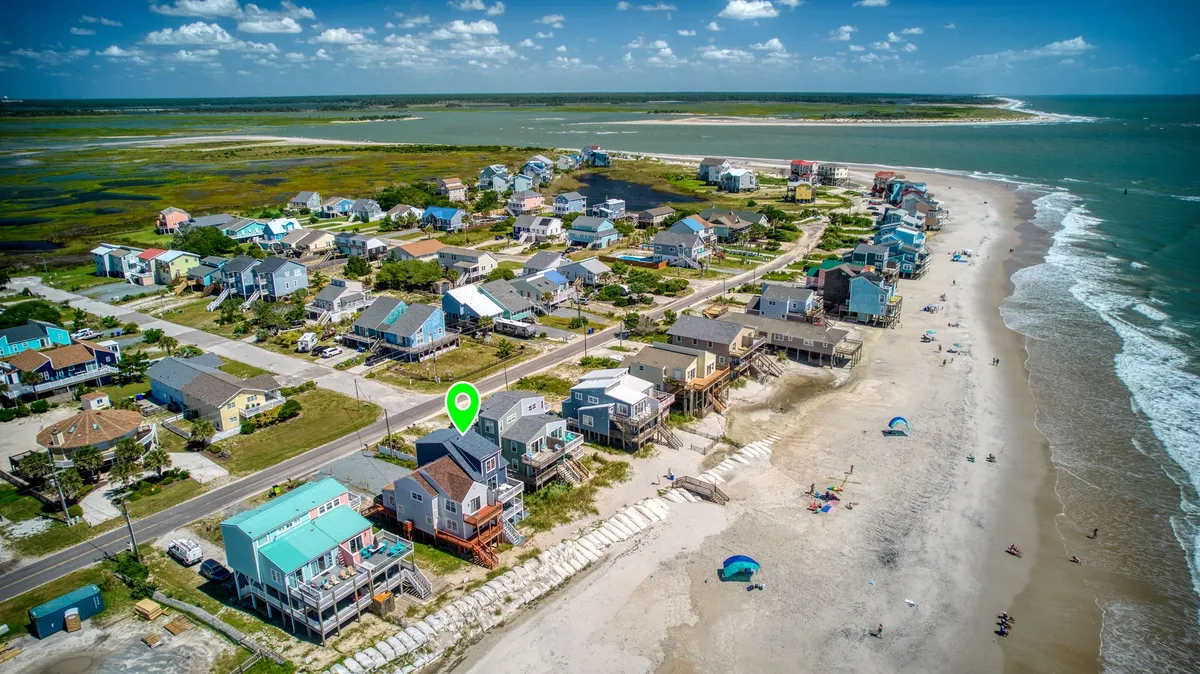 A wide-angle perspective of beachfront houses facing an expansive, sandy beach with rolling ocean waves and lush greenery at the edges. The inviting, unspoiled shoreline stretches into the distance, perfect for walks, shell-collecting, and family play.