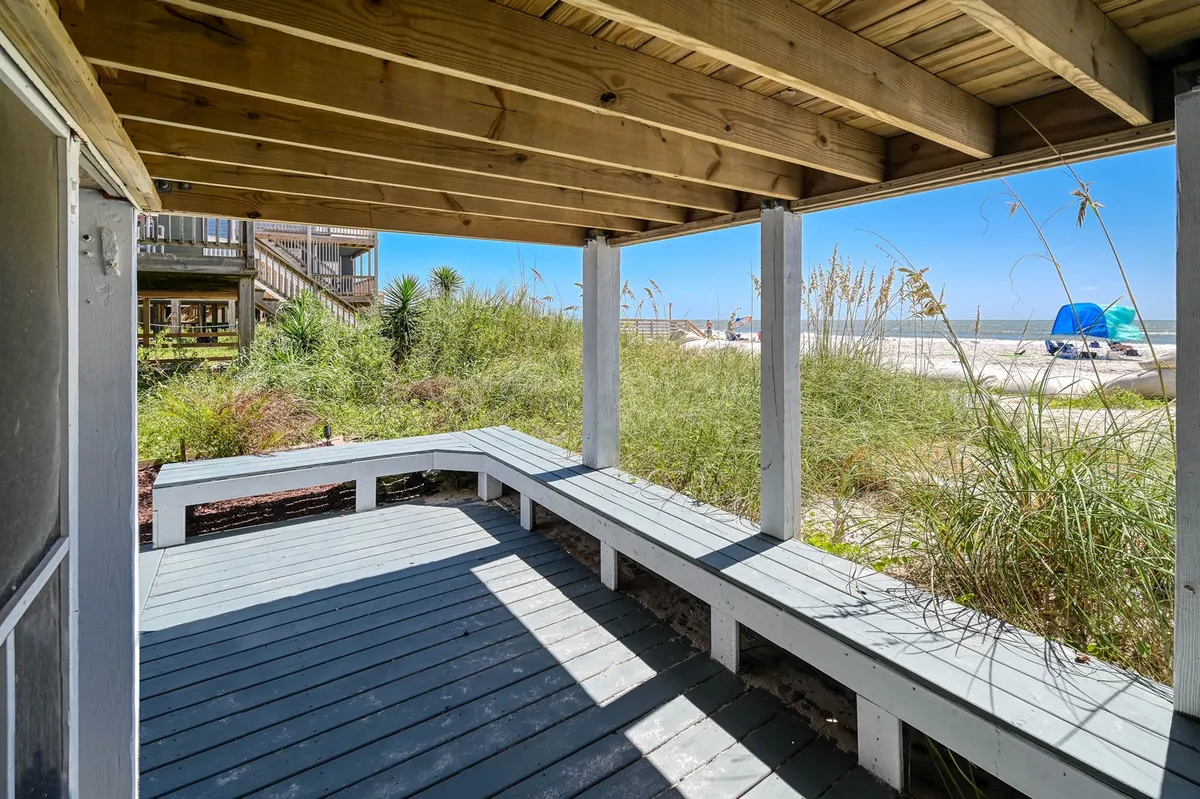A peaceful wooden seating area on a deck overlooking the beach, surrounded by greenery and native plants. The ocean is clearly visible beyond the deck, creating a serene setting for relaxation. The scene is tranquil, evoking the sound of waves and the feel of a soft breeze.
