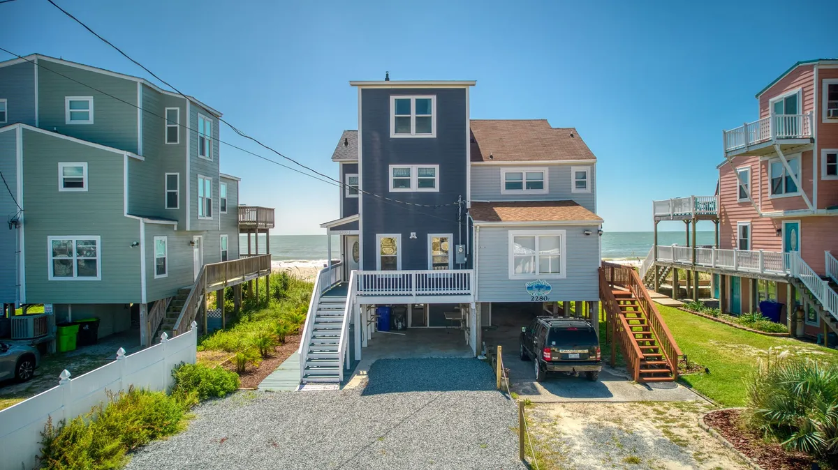 The image showcases an inviting beach house with a welcoming facade, set directly along the pristine sands of North Topsail Beach. The home stands out with its expansive deck facing the ocean, neighboring houses nearby, and a clear, sunny sky that hints at beautiful weather. The setting evokes easy access to the beach, relaxation on the porch, and memorable family gatherings overlooking the waves.