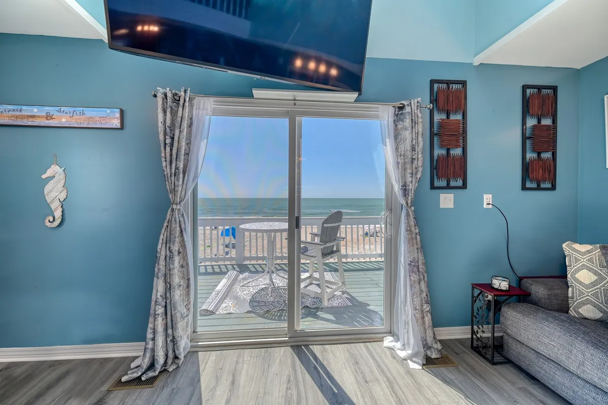 A cozy living space with a sofa positioned to take in the beach view through large sliding glass doors. The airy, light-filled room draws the outdoors in, seamlessly blending interior comfort and spectacular natural scenery. Thoughtful decor and wall art enhance the relaxing vibe.