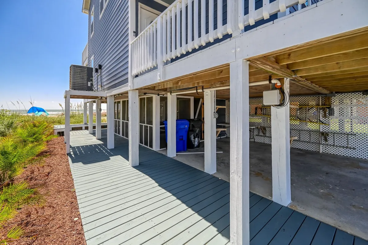 A practical and inviting view underneath the house, showing decking, outdoor furniture, and a blue recycling bin. The shaded space is perfect for cleaning up after the beach or storing gear, and is adjacent to the beach access point.