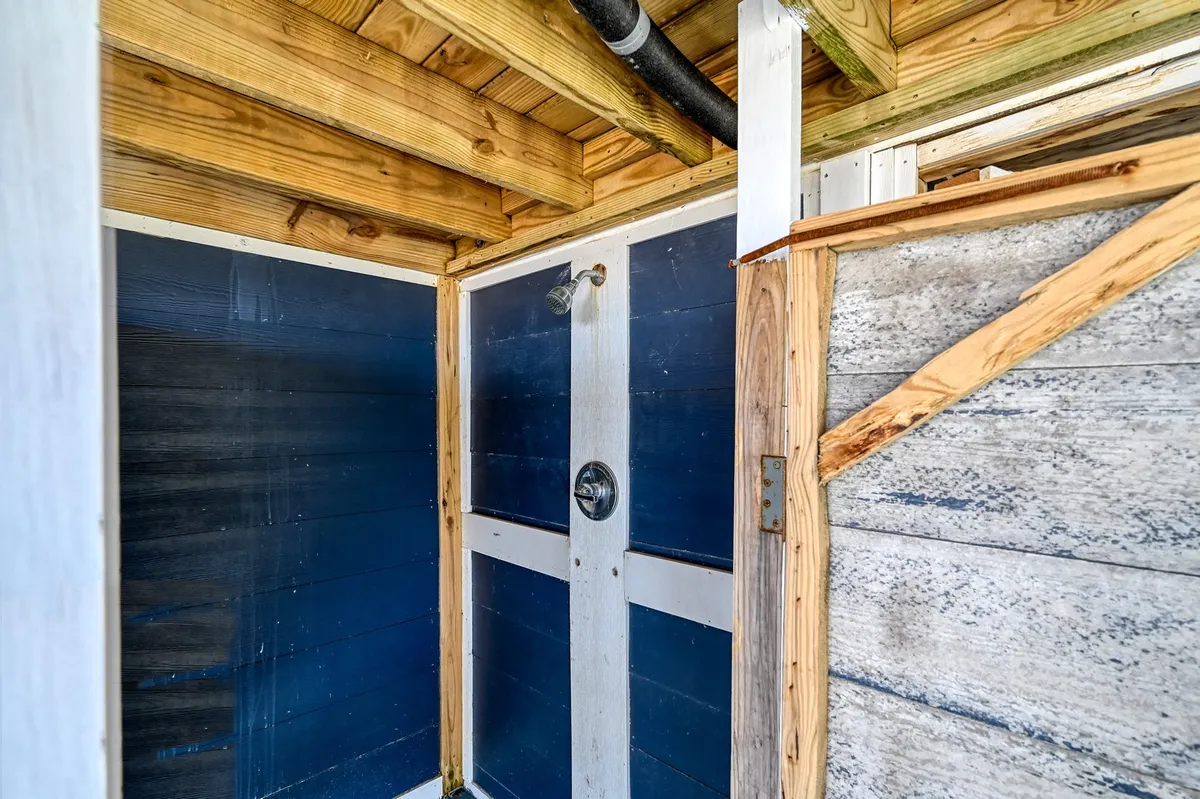 A refreshing outdoor shower setup with a prominent showerhead, rustic wooden beams, and blue-painted walls, designed for rinsing off after sandy beach days. The shower is airy and open, evoking a sense of nature and simplicity.