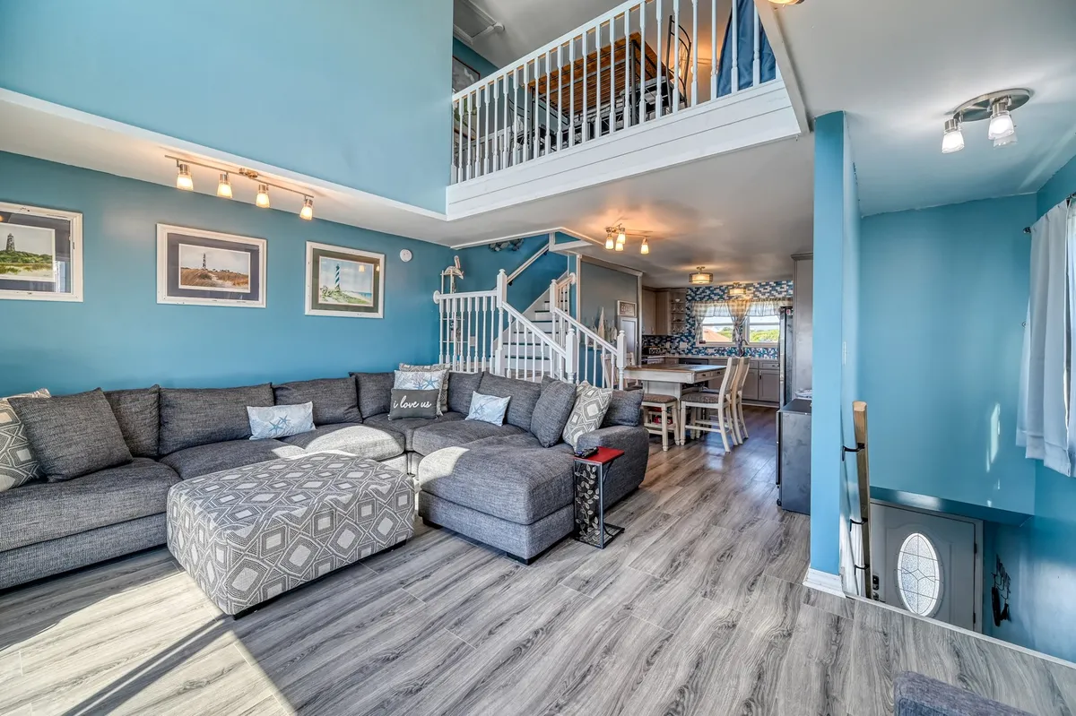 This image features a spacious open-plan living area with a large sectional sofa, a dining table, and a visible staircase. The room is filled with natural light, creating a welcoming atmosphere that's ideal for family gatherings, movie nights, and shared meals. The design suggests openness and connection, with areas for both relaxation and socializing.