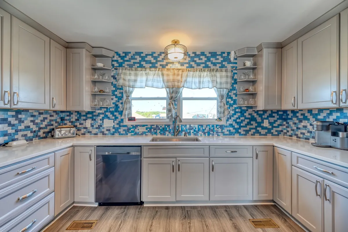 The kitchen area is highlighted here, featuring a colorful backsplash, clean quartz countertops, and modern lighting. The scene is lively, suggesting meal preparation, family baking sessions, or hosting brunches. A window with curtains brings in daylight, and the space feels both modern and homey.