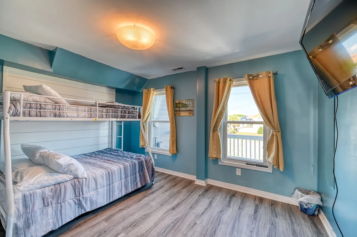 A comfortable bunk room featuring a bunk bed, large windows with flowing curtains, and a TV mounted on the wall. The atmosphere is casual and kid-friendly, ideal for sleeping, relaxing, or watching TV together.