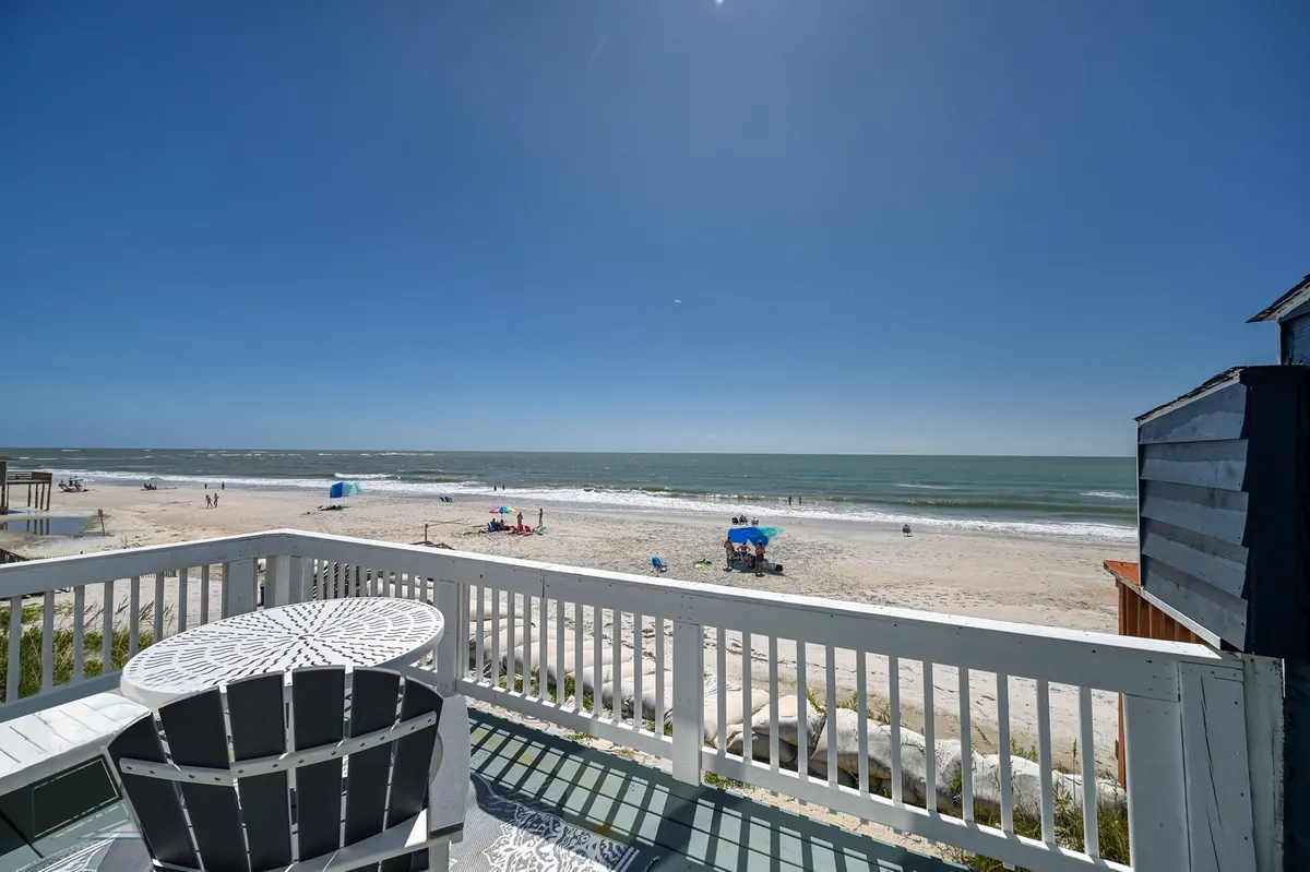 A sweeping view of the sandy beach and ocean with outdoor seating and umbrellas set up for relaxation. The scene looks inviting, capturing the essence of early morning walks, playful moments on the sand, and the expansive shoreline just steps from the property.