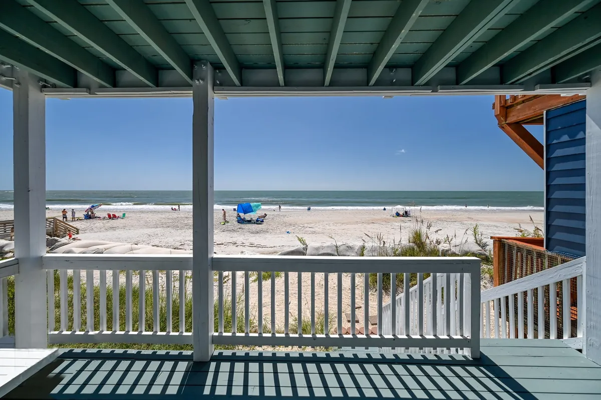 A lively, inviting beach scene featuring the sparkling ocean, golden sands, people sunbathing, and colorful umbrellas dotting the shoreline. Families and children are visible enjoying classic beach activities like swimming, playing volleyball, and building sandcastles, with the boardwalk and nearby homes subtly in view.
